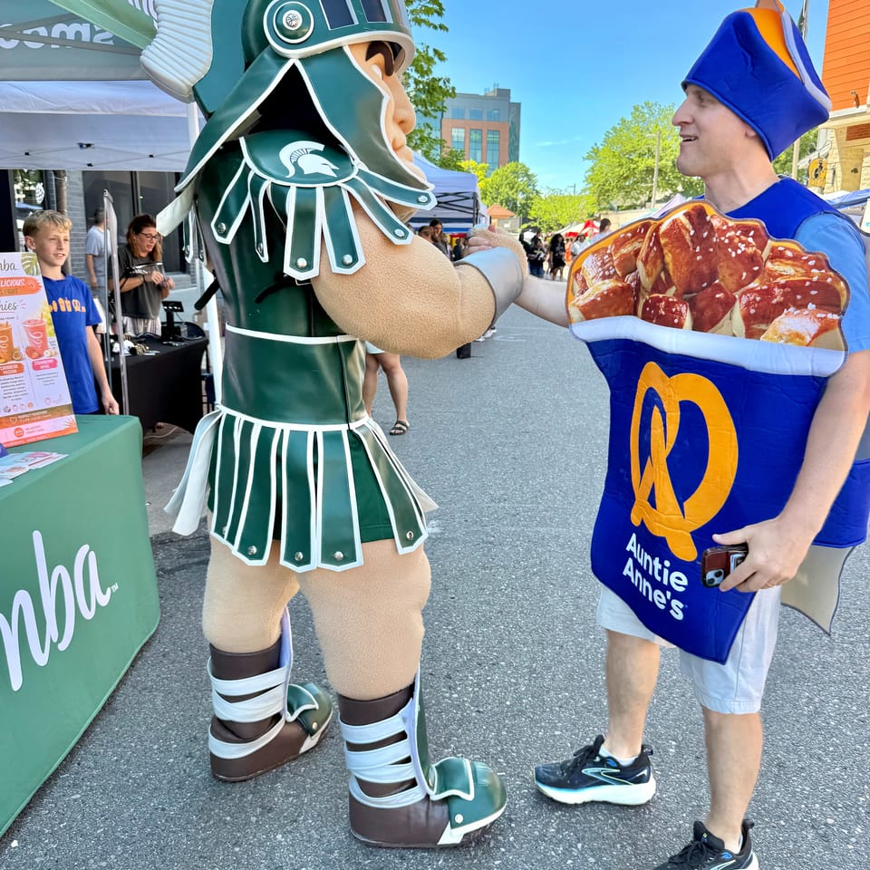 A Spartan mascot shakes hands with a person in an Auntie Anne's pretzel costume outdoors.