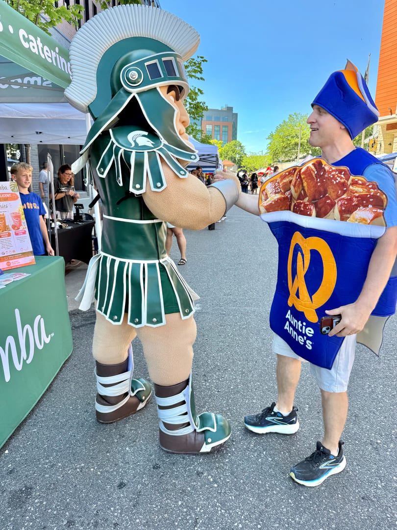 A Spartan mascot shakes hands with a person in an Auntie Anne's pretzel costume outdoors.