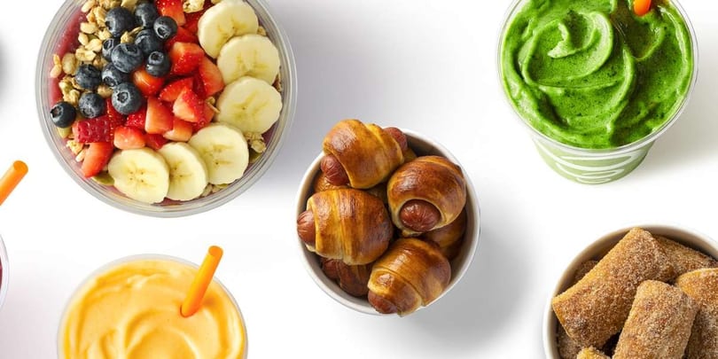 Overhead view of a Jamba açaí bowl, green smoothie, orange smoothie, pretzel dogs, and churro bites.